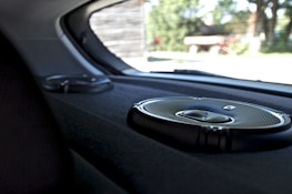 A close-up view of the interior of a car showing two circular audio speakers installed on the rear deck. The larger speaker is in the foreground, displaying a metallic grille and sleek design. Outside the car, blurry greenery and structures are visible through the rear window.