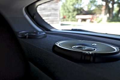 A close-up view of the interior of a car showing two circular audio speakers installed on the rear deck. The larger speaker is in the foreground, displaying a metallic grille and sleek design. Outside the car, blurry greenery and structures are visible through the rear window.