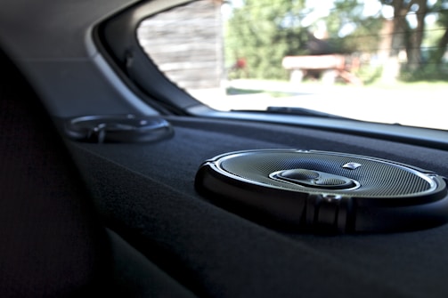 A close-up view of the interior of a car showing two circular audio speakers installed on the rear deck. The larger speaker is in the foreground, displaying a metallic grille and sleek design. Outside the car, blurry greenery and structures are visible through the rear window.