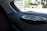 A close-up view of the interior of a car showing two circular audio speakers installed on the rear deck. The larger speaker is in the foreground, displaying a metallic grille and sleek design. Outside the car, blurry greenery and structures are visible through the rear window.