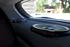 A close-up view of the interior of a car showing two circular audio speakers installed on the rear deck. The larger speaker is in the foreground, displaying a metallic grille and sleek design. Outside the car, blurry greenery and structures are visible through the rear window.