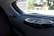 A close-up view of the interior of a car showing two circular audio speakers installed on the rear deck. The larger speaker is in the foreground, displaying a metallic grille and sleek design. Outside the car, blurry greenery and structures are visible through the rear window.