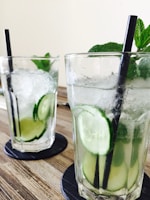 Two glasses of refreshing beverages, garnished with slices of cucumber and fresh mint leaves, are placed on black coasters. The drinks contain ice and are served with black straws. They are set on a wooden table surface.