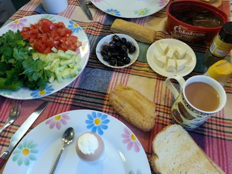 A colorful breakfast spread on a plaid tablecloth including fresh vegetables like tomatoes, cucumber, and parsley on a floral plate, a boiled egg in an egg cup, sliced cheese, black olives, and a variety of bread. There's also a cup of tea or coffee, a container of chocolate spread, a spice bottle, and a honey jar.