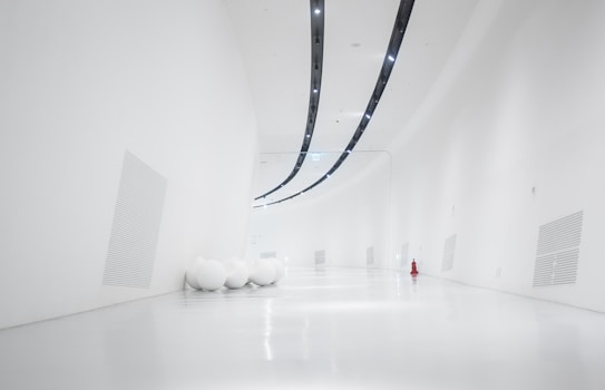 A minimalistic and modern interior space with smooth, curved white walls and ceiling. The floor is also white and shiny, creating a seamless and airy environment. There are a few large, round white spheres scattered to the left side and a bright red fire hydrant on the right, adding a pop of color.