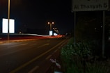 A nighttime street scene with long exposure light trails from vehicles. The road curves to the left, lined with streetlights that create a warm glow. On the right side, there's vegetation and a street sign with 'Al Thanyah 5' visible. Bright, illuminated billboards stand out in the background, adding contrast to the dark surroundings.