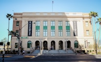 The image depicts a large, historic building with a beige facade, green window frames, and an American flag on top. The building is identified as the Mob Museum by large banners on its sides. There are wide steps leading up to the entrance and several people can be seen walking near the entrance. Palm trees are present around the building, and a street sign indicates the location is Stewart Avenue.