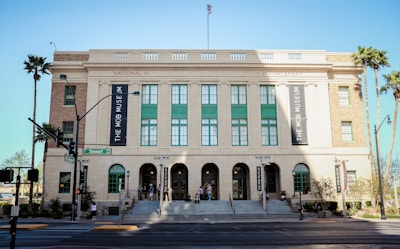 The image depicts a large, historic building with a beige facade, green window frames, and an American flag on top. The building is identified as the Mob Museum by large banners on its sides. There are wide steps leading up to the entrance and several people can be seen walking near the entrance. Palm trees are present around the building, and a street sign indicates the location is Stewart Avenue.