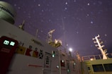 A ship's deck is visible at night under a starry sky. Various navigational and communication structures, as well as signal flags, are present on the vessel. The moon is partially visible, casting a soft light on the scene, while the colors and shapes of lights from equipment and signs can be seen.