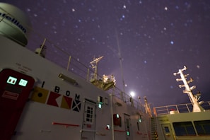 A ship's deck is visible at night under a starry sky. Various navigational and communication structures, as well as signal flags, are present on the vessel. The moon is partially visible, casting a soft light on the scene, while the colors and shapes of lights from equipment and signs can be seen.