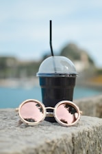A black plastic cup with a dome lid and straw is placed on a stone surface. Beside it, a pair of round sunglasses with reflective pink lenses rests, mirroring the surrounding landscape.