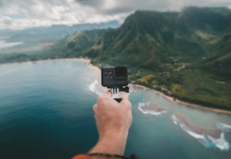A hand holding a camera extends over a scenic view of a coastline with lush green mountains and a stretch of ocean. The landscape features rolling hills covered in greenery and clouds hanging low in the sky, creating a dramatic backdrop.