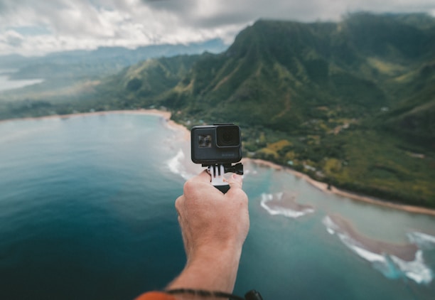 A hand holding a camera extends over a scenic view of a coastline with lush green mountains and a stretch of ocean. The landscape features rolling hills covered in greenery and clouds hanging low in the sky, creating a dramatic backdrop.