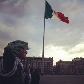 A soldier stands saluting in foreground, wearing a green uniform with white details, facing a large Mexican flag on a pole. Surrounding the scene are historic buildings with a cloudy sky adding a dramatic atmosphere.
