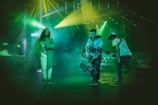 Three individuals are present in a dimly lit environment with vibrant lighting effects. One person, wearing a long coat and light pants, stands on the left side, while two others are on the right, handling film equipment. The atmospheric green and yellow lights and smoky background give the scene a cinematic and dynamic feel.