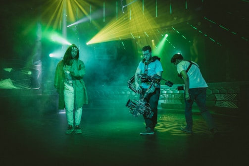 Three individuals are present in a dimly lit environment with vibrant lighting effects. One person, wearing a long coat and light pants, stands on the left side, while two others are on the right, handling film equipment. The atmospheric green and yellow lights and smoky background give the scene a cinematic and dynamic feel.