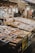 An outdoor market stall featuring a variety of packaged goods and seafood. The packages are displayed on tables made of stacked crates under hanging light bulbs. Surrounding the display, there are cardboard boxes and handwritten labels. The setting has a rustic and bustling market atmosphere.