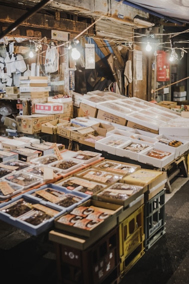 An outdoor market stall featuring a variety of packaged goods and seafood. The packages are displayed on tables made of stacked crates under hanging light bulbs. Surrounding the display, there are cardboard boxes and handwritten labels. The setting has a rustic and bustling market atmosphere.