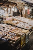 An outdoor market stall featuring a variety of packaged goods and seafood. The packages are displayed on tables made of stacked crates under hanging light bulbs. Surrounding the display, there are cardboard boxes and handwritten labels. The setting has a rustic and bustling market atmosphere.