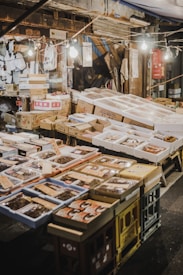 An outdoor market stall featuring a variety of packaged goods and seafood. The packages are displayed on tables made of stacked crates under hanging light bulbs. Surrounding the display, there are cardboard boxes and handwritten labels. The setting has a rustic and bustling market atmosphere.