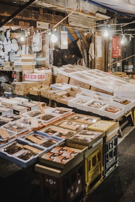 An outdoor market stall featuring a variety of packaged goods and seafood. The packages are displayed on tables made of stacked crates under hanging light bulbs. Surrounding the display, there are cardboard boxes and handwritten labels. The setting has a rustic and bustling market atmosphere.