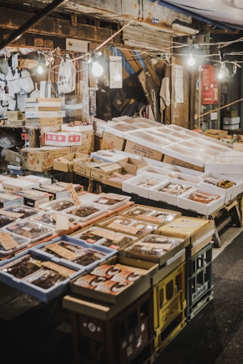 An outdoor market stall featuring a variety of packaged goods and seafood. The packages are displayed on tables made of stacked crates under hanging light bulbs. Surrounding the display, there are cardboard boxes and handwritten labels. The setting has a rustic and bustling market atmosphere.