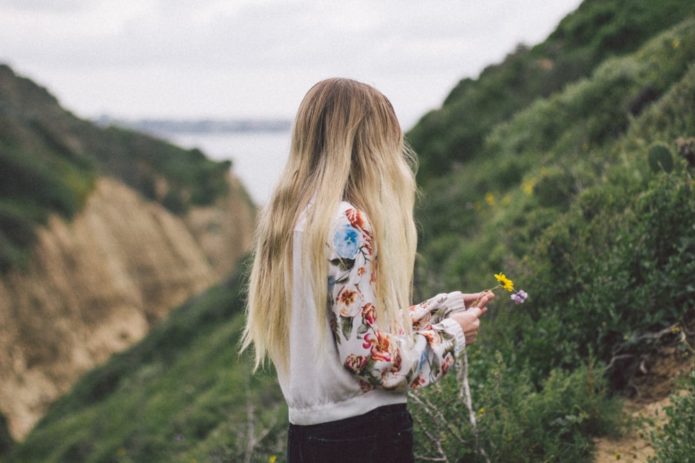 흰색, 녹색, 빨간색 꽃무늬 긴팔 셔츠를 입은 여자가 낮 동안 녹색 잎 식물로 덮인 언덕에 서 있습니다.