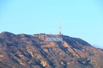 A famous hillside landmark is visible under a clear blue sky. An iconic sign with large white letters is prominently displayed on the rugged terrain of the hill. Near the top, a tall radio tower stands against the backdrop of the sky.