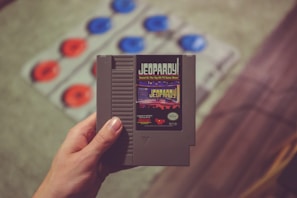 Close-up of hands holding a well-loved retro game cartridge against a backdrop of arcade machines.