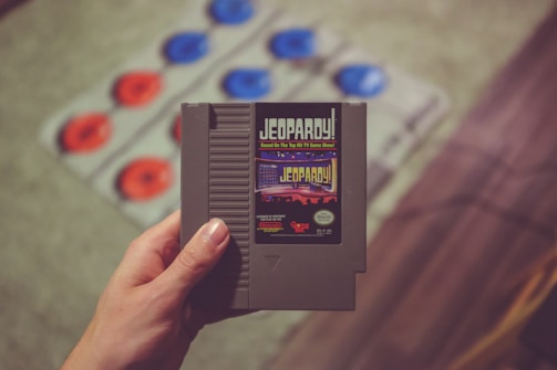 A hand is holding a vintage Jeopardy! video game cartridge. The background features a blurred out detail of a game mat with red and blue circles.
