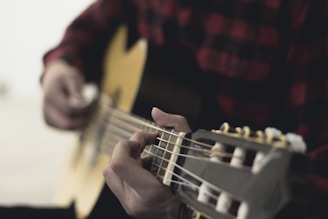 A person is playing an acoustic guitar, focusing on the fingers pressing the strings. The background is softly blurred, highlighting the action of strumming.
