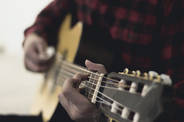A person is playing an acoustic guitar, focusing on the fingers pressing the strings. The background is softly blurred, highlighting the action of strumming.