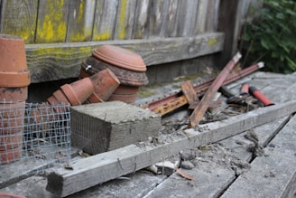 An assortment of gardening and construction materials is scattered on a wooden surface. Several terracotta pots are stacked alongside pieces of wood and metal, including rusty rods and a wire mesh. A pair of red-handled garden shears is visible, surrounded by debris and dirt. The background features a wooden fence with patches of yellow-green moss.