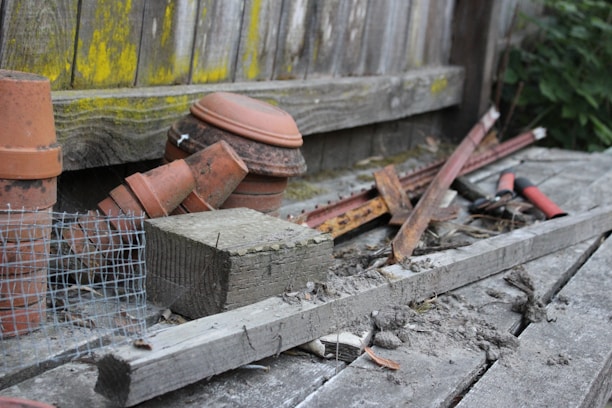 An assortment of gardening and construction materials is scattered on a wooden surface. Several terracotta pots are stacked alongside pieces of wood and metal, including rusty rods and a wire mesh. A pair of red-handled garden shears is visible, surrounded by debris and dirt. The background features a wooden fence with patches of yellow-green moss.