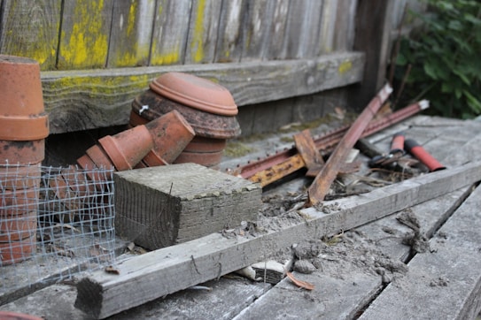An assortment of gardening and construction materials is scattered on a wooden surface. Several terracotta pots are stacked alongside pieces of wood and metal, including rusty rods and a wire mesh. A pair of red-handled garden shears is visible, surrounded by debris and dirt. The background features a wooden fence with patches of yellow-green moss.