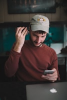 A young man with a beard and a casual cap is engaged in a task involving two smartphones. He wears a maroon sweatshirt and sits at a desk with a laptop. The room's dim lighting, coupled with his focused expression, suggests concentration.