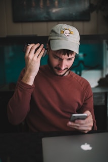 A young man with a beard and a casual cap is engaged in a task involving two smartphones. He wears a maroon sweatshirt and sits at a desk with a laptop. The room's dim lighting, coupled with his focused expression, suggests concentration.