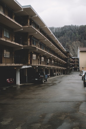 A multi-story motel with rows of balconies lined up along the building. Several parked cars are visible in the parking area beneath the structure, with a backdrop of forested hills and overcast sky.