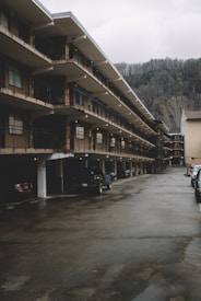 A multi-story motel with rows of balconies lined up along the building. Several parked cars are visible in the parking area beneath the structure, with a backdrop of forested hills and overcast sky.