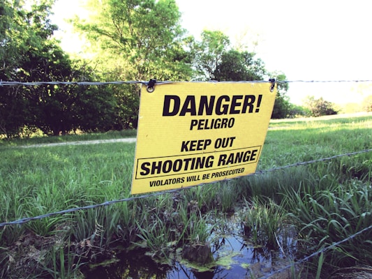 A yellow warning sign is attached to a barbed wire fence, set in front of a backdrop of lush greenery. The sign reads 'Danger! Peligro Keep Out Shooting Range Violators Will Be Prosecuted' in bold black letters.