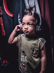 A child wearing headphones stands in a room, holding the headphones close to one ear. The child is dressed in a green shirt with cartoon graphics and camo design elements. In the background, various pieces of clothing hang on a rack, including a sports jersey with red and black colors.