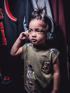 A child wearing headphones stands in a room, holding the headphones close to one ear. The child is dressed in a green shirt with cartoon graphics and camo design elements. In the background, various pieces of clothing hang on a rack, including a sports jersey with red and black colors.