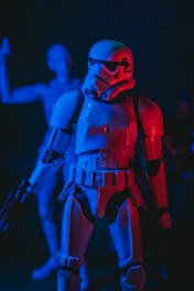 A plastic figure of a stormtrooper is prominently featured, illuminated by intense blue and red lighting. The stormtrooper is holding a blaster and has a futuristic, armored appearance. In the background, another out-of-focus figure adds depth to the scene.
