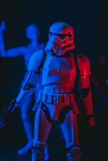 A plastic figure of a stormtrooper is prominently featured, illuminated by intense blue and red lighting. The stormtrooper is holding a blaster and has a futuristic, armored appearance. In the background, another out-of-focus figure adds depth to the scene.