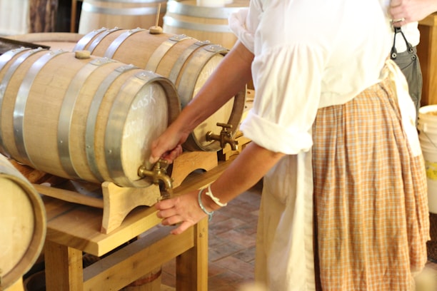 A person in period-style clothing is operating a tap on a wooden cask in a rustic setting. The barrels are set up on wooden racks and the environment suggests a historical or artisanal atmosphere.