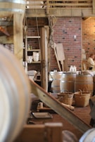 A rustic interior with wooden beams and brick walls. There are large wooden barrels and wicker baskets spread around the space. Various kitchen items like plates and mugs are visible in the background. A rope hangs from the structure above, adding to the traditional look.