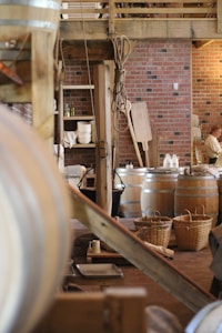 A rustic interior with wooden beams and brick walls. There are large wooden barrels and wicker baskets spread around the space. Various kitchen items like plates and mugs are visible in the background. A rope hangs from the structure above, adding to the traditional look.