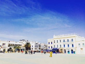 A large open plaza is surrounded by white buildings with blue accents under a bright blue sky. People are scattered throughout the plaza, some walking, others gathered in small groups. Palms and small carts add to the lively scene.