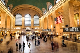 A grand hall with an ornate ceiling, large arched windows, and a prominent American flag. People are walking around the spacious area, and information boards are visible on the walls. The arched design and lighting create a majestic atmosphere.