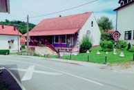 A quaint house with a red-tiled roof and white walls, featuring purple accents and a painted mural on the exterior. The house is adorned with hanging flower pots and surrounded by lush greenery and manicured lawns. The front yard has several decorative items resembling chickens and a small shrine or decorative structure on the side. The road in front of the house shows a large white arrow, and other houses are visible in the background, indicating a residential area.